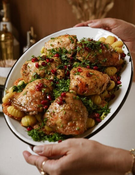 A person holds an oval platter filled with roasted chicken thighs, garnished with fresh herbs and pomegranate seeds, served with sheet pan gnocchi. The dish is set against a kitchen backdrop.