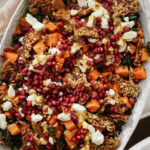 A close-up of a colorful salad featuring sweet potatoes, crumbled feta, pomegranate seeds, kale, and crunchy seeds or nuts, all drizzled with a light dressing. The dish is served in an oval platter.
