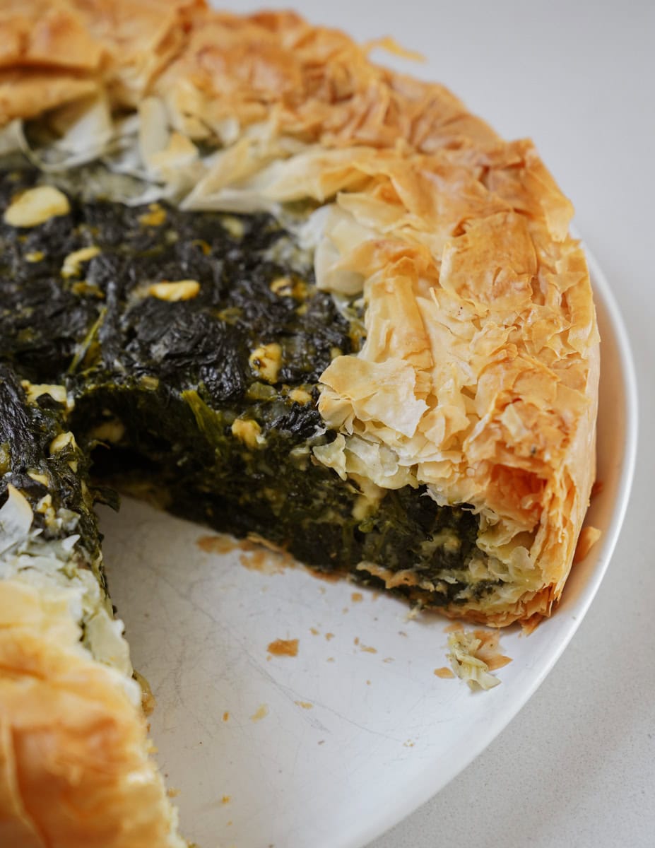 A close-up of a spinach and cheese pie with flaky golden-brown crust on a white plate. A slice has been removed, revealing the dark green spinach filling mixed with cheese.