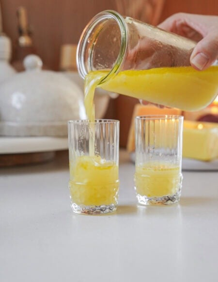 A hand pours fresh orange juice from a glass pitcher into one of two clear, textured glasses on a white countertop. The background features a blurred kitchen setting with various items.