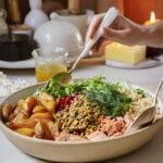A hand with a fork reaches into a large bowl filled with a variety of colorful ingredients: roasted potatoes, pistachios, pomegranate seeds, greens, couscous, dill, and shredded salmon. In the background are condiments and a lit candle.