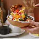 A person holds a ceramic bowl filled with a colorful salad of cherry tomatoes, lentils, and herbs. Theyre using a spoon to serve more salad into the bowl. A tray of ingredients and kitchen items are blurred in the background.