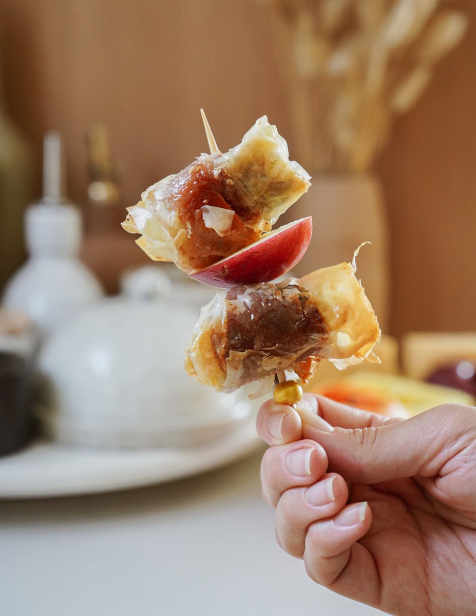 A hand holds a skewer with two pieces of crumpled, golden-brown pastry and a slice of red fruit. A blurred background shows kitchen items and soft lighting.