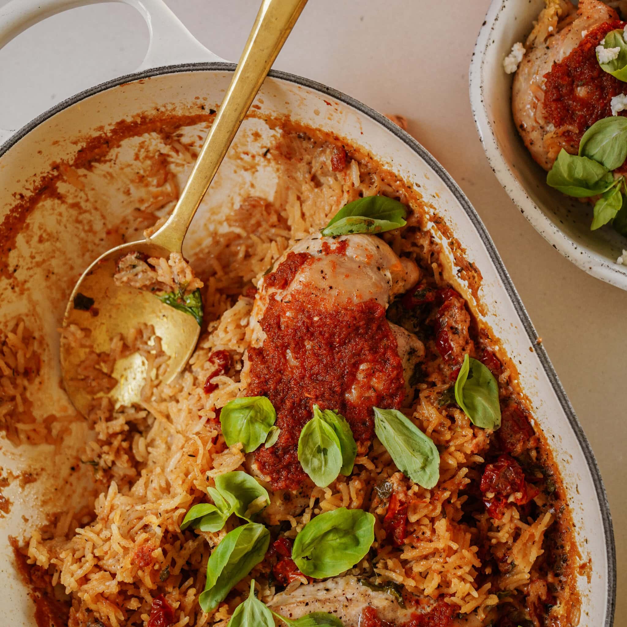 A baked dish of seasoned chicken thighs and rice garnished with fresh basil leaves, in a white oval dish. A golden spoon is placed on the left side. A small bowl with a portion of the dish is visible in the background.