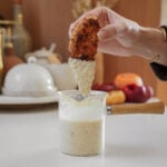 A hand is dipping a piece of breaded and fried food into a small glass pot filled with a creamy sauce. The pot has a wooden handle and is on a kitchen counter with blurred kitchen items in the background.
