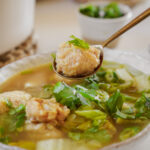 A spoon holding matzo ball above a bowl of matzo ball soup, garnished with fresh cilantro. Background shows a small bowl with more cilantro.