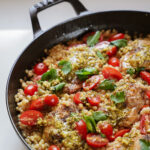A skillet filled with pearl couscous, cherry tomatoes, and cooked chicken thighs topped with green pesto. Fresh parsley leaves are scattered on top for garnish.