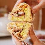 Two hands hold a halved sandwich with a crispy tortilla shell, filled with scrambled eggs, chicken, and herbs. The filling is visible and neatly packed, with a blurry background of a table setting.