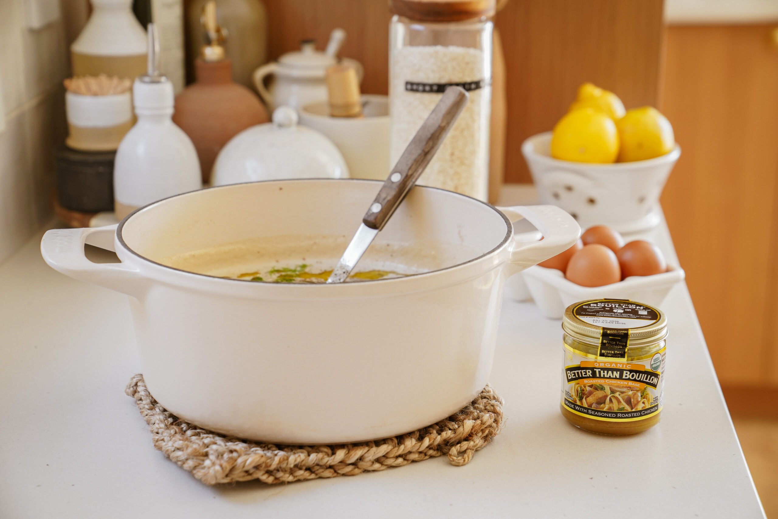 A white pot with a spoon rests on a kitchen counter, surrounded by ingredients including eggs, lemons, a jar labeled Better Than Bouillon, and a container of rice. Decorative pottery and kitchen items are in the background.