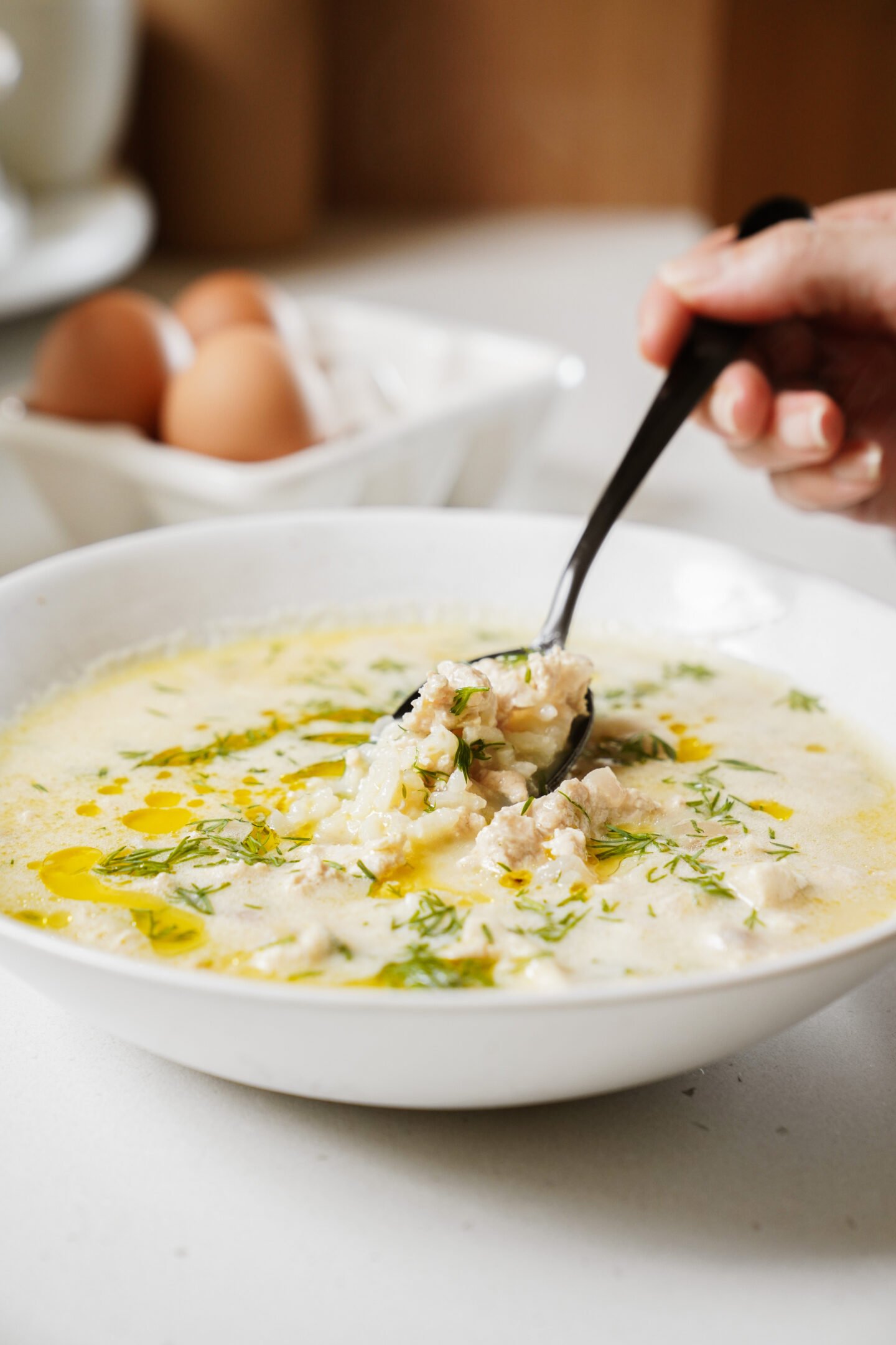 A person holds a spoon filled with creamy soup from a white bowl. The soup is garnished with herbs and drizzled with oil. In the background, a container with brown eggs is visible on a white countertop.