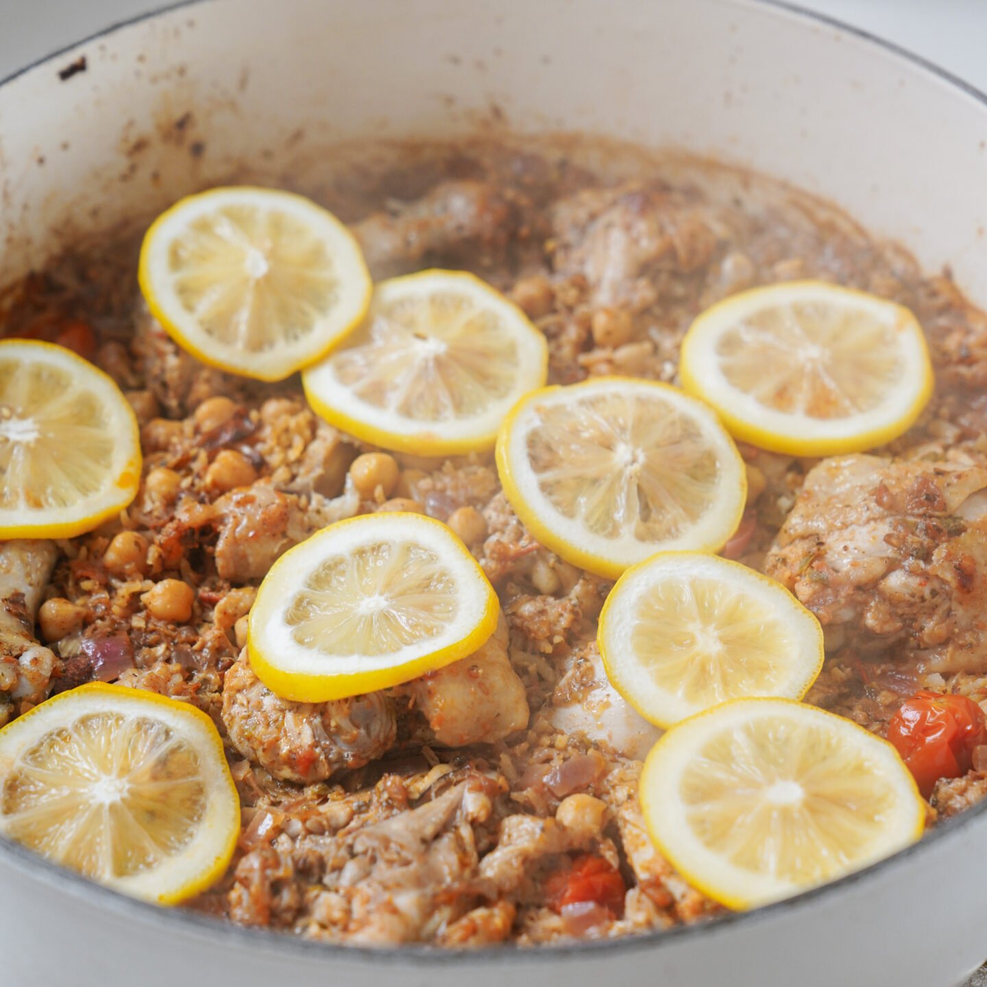 A pot containing cooked chicken pieces mixed with chickpeas, tomatoes, and spices, topped with slices of lemon. The dish appears to be a hearty stew or casserole, with visible steam, suggesting it is freshly cooked.