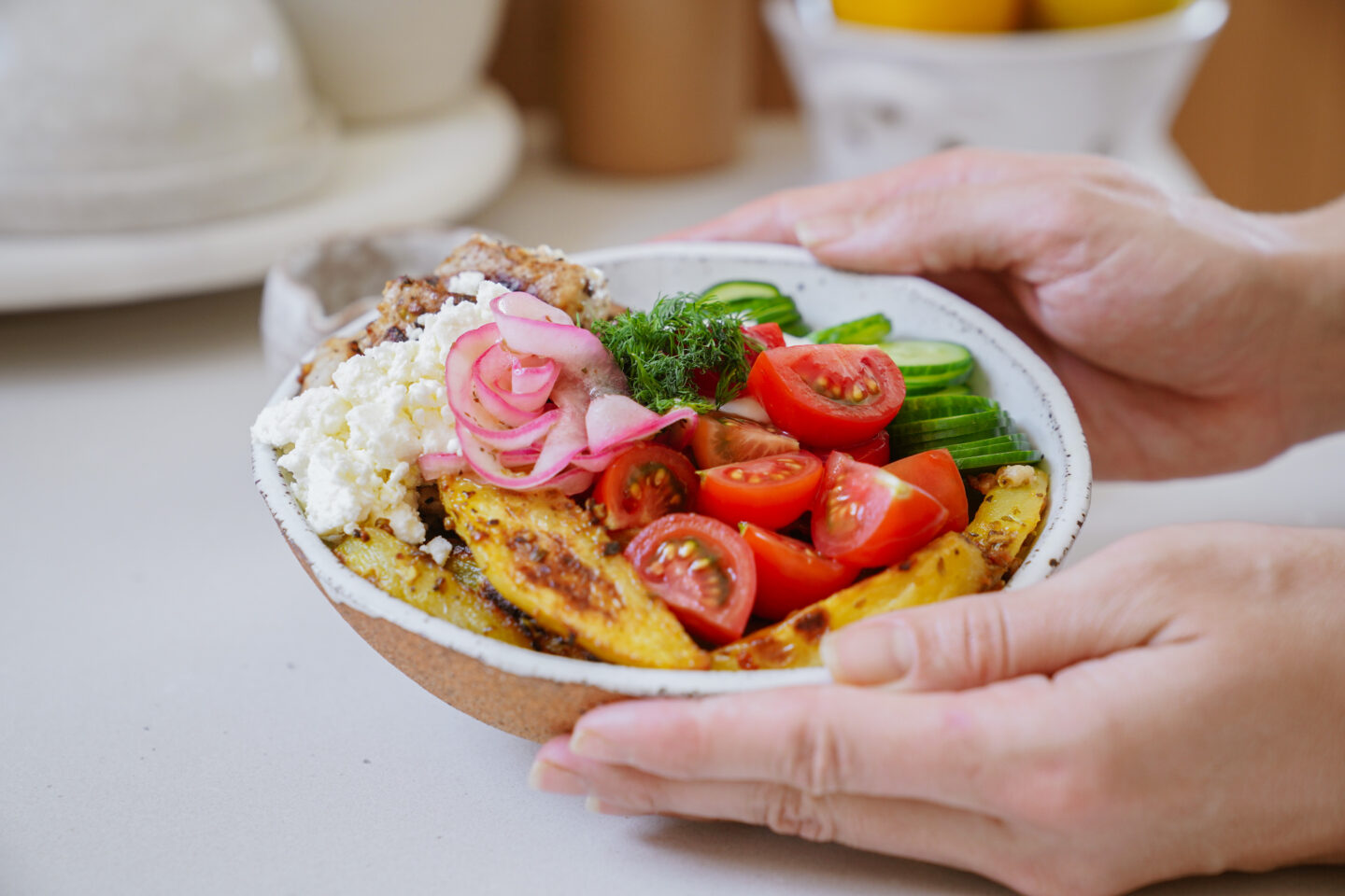 A person holds a bowl filled with roasted potatoes, cherry tomatoes, cucumber slices, pickled onions, dill, feta cheese, and grains. The colorful ingredients are arranged neatly, creating a vibrant and appetizing presentation.