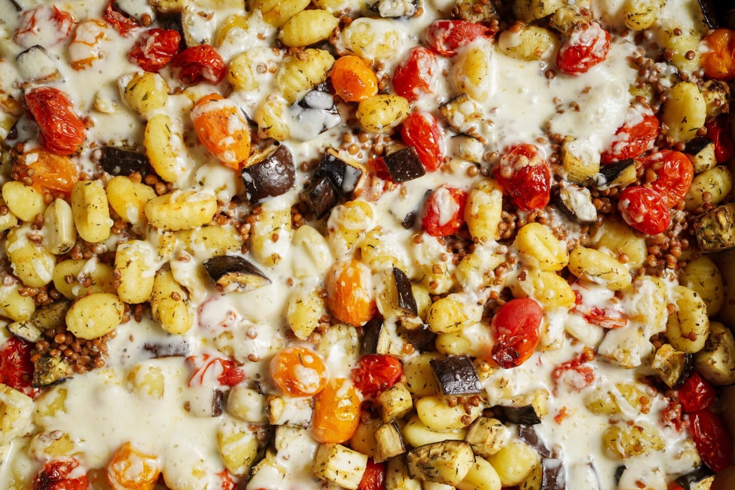 A close-up of a baked gnocchi dish featuring golden gnocchi pieces, cherry tomatoes, eggplant chunks, and melted cheese. The ingredients are intermingled, creating a colorful and appetizing texture.