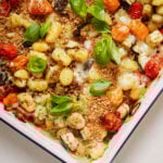 A baked gnocchi dish topped with melted cheese, roasted cherry tomatoes, eggplant, and zucchini. Fresh basil leaves garnish the top, adding color. The edges of the dish are slightly golden, indicating a crisp finish.