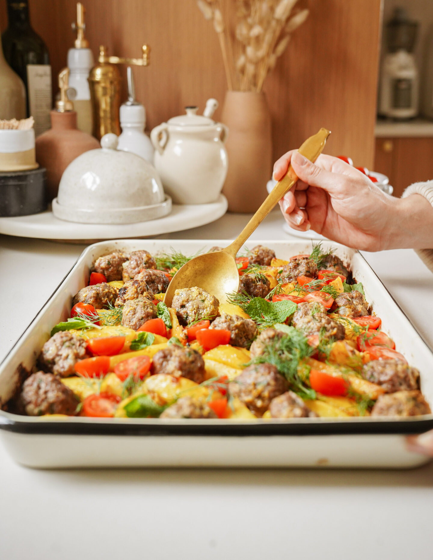 A person serves a tray of roasted meatballs with colorful vegetables like carrots and zucchini using a gold spoon. The dish sits on a white countertop with kitchen items and decor in the background.