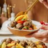 A hand holds a rustic bowl filled with seasoned meatballs, roasted potato wedges, and cherry tomatoes, garnished with fresh herbs. A golden spoon is scooping the food. Background features blurred kitchen items.