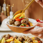 A hand holds a rustic bowl filled with seasoned meatballs, roasted potato wedges, and cherry tomatoes, garnished with fresh herbs. A golden spoon is scooping the food. Background features blurred kitchen items.