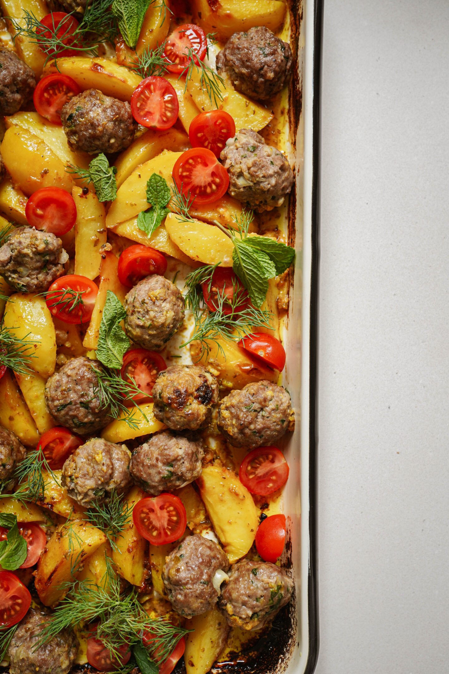 A baked dish featuring meatballs, sliced yellow potatoes, cherry tomato halves, and fresh herbs like dill and mint on a sheet pan. The ingredients are evenly spread, with golden-brown edges visible on the pan.