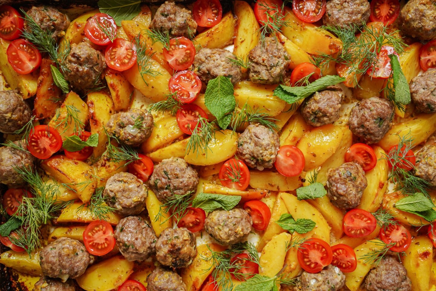 A tray of baked potatoes and meatballs garnished with sliced cherry tomatoes, dill, and mint leaves. The dish features a colorful mix of golden potatoes and appetizing meatballs arranged on a textured surface.
