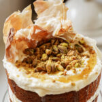 A close-up of a layered cake topped with whipped cream, a drizzle of honey, crumbled pistachios, and crispy phyllo pastry pieces, creating a textured and inviting appearance.