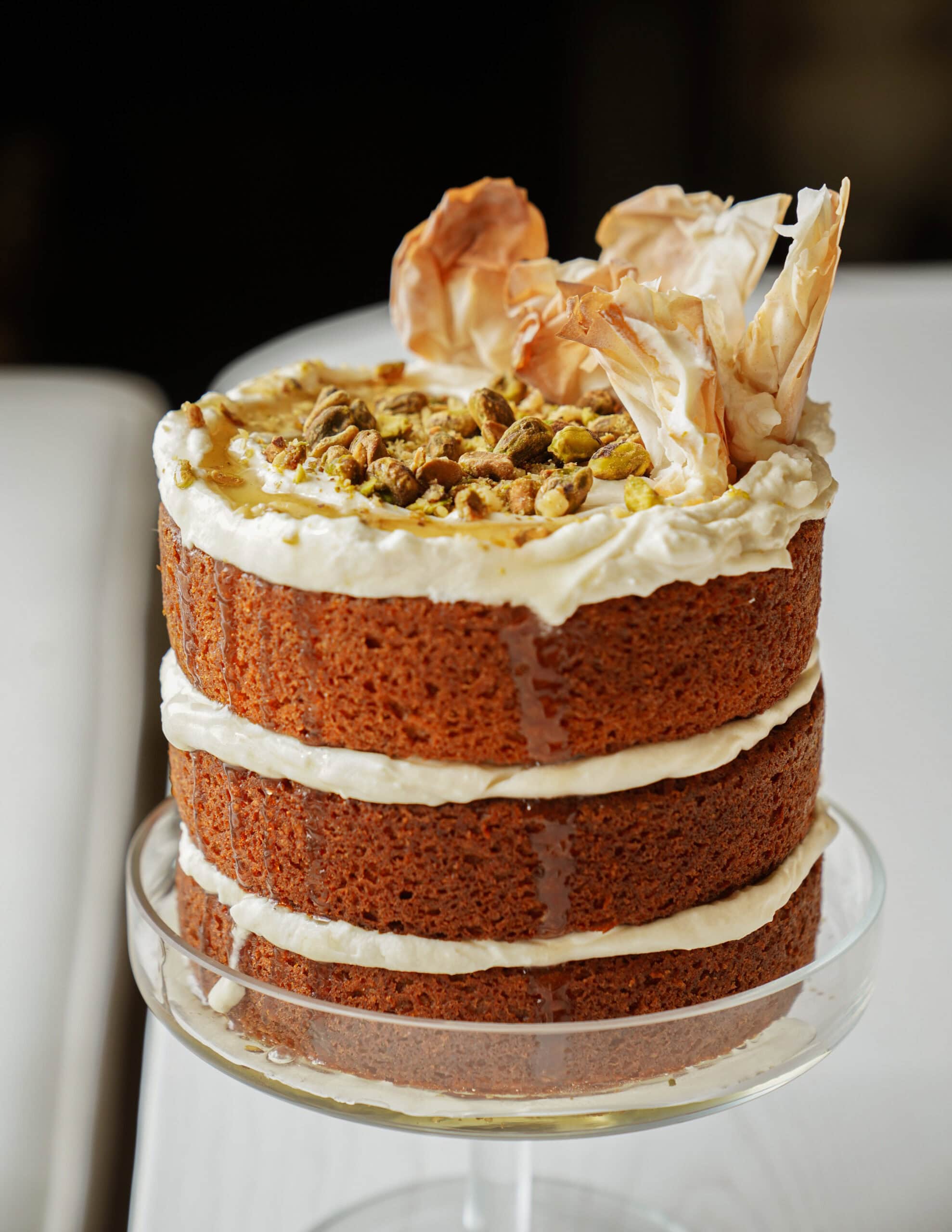 Three-layer naked cake topped with cream cheese frosting, chopped pistachios, and decorative pastry flakes, displayed on a glass cake stand. The cakes layers are visible with white frosting in between.