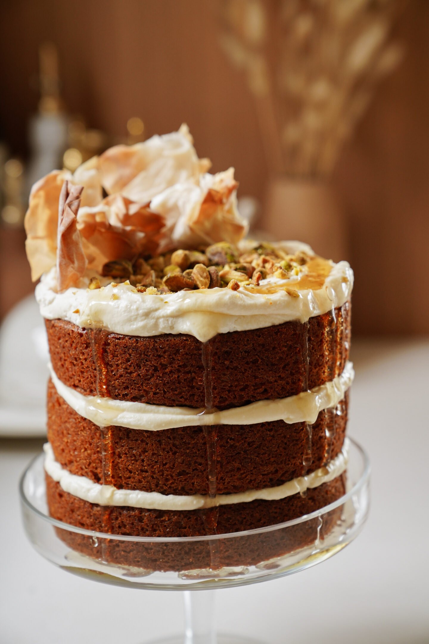 Three-layered naked cake on a glass stand, with cream filling and topped with pistachios and decorative dried leaves, set against a soft-focus background of blurred warm-toned decor.