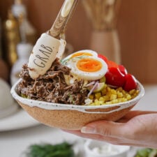 A hand holds a bowl filled with shredded beef, sliced boiled egg, diced potatoes, cherry tomatoes, red onions, and greens. A spatula with GRAB written on it tops the bowl with a dollop of creamy sauce.