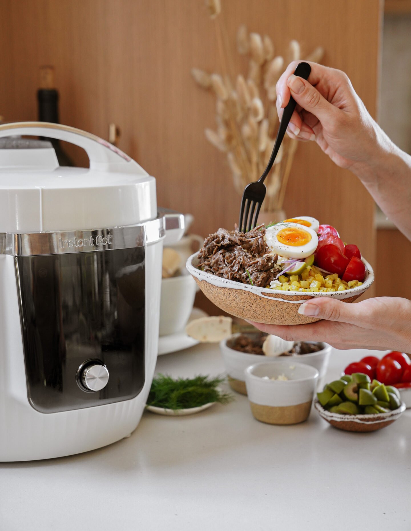 A person holds a bowl of shredded meat, boiled egg, cherry tomatoes, and corn, using a fork. A white Instant Pot sits on the counter nearby with ingredients and garnishes in the background.
