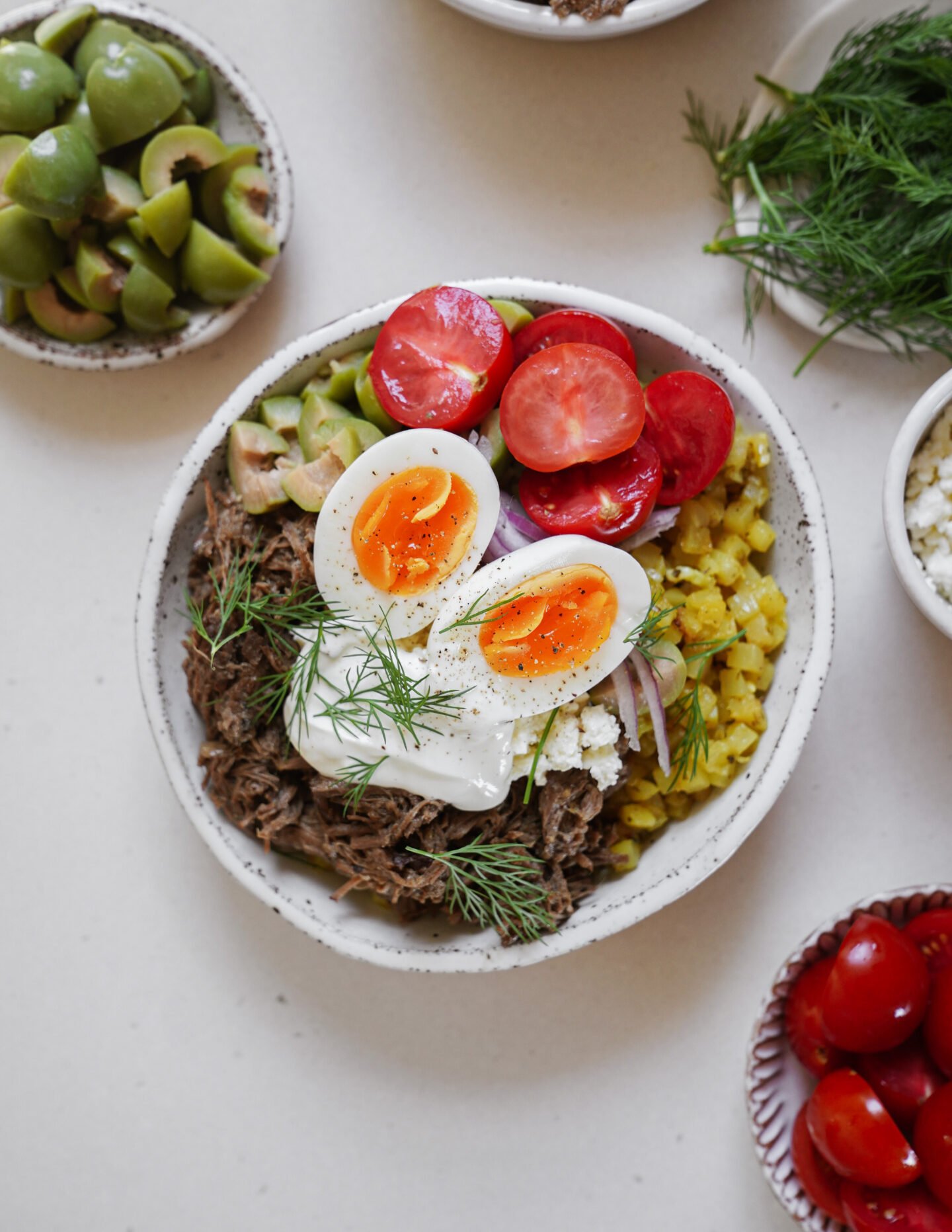 A bowl filled with shredded meat, sliced green olives, yellow rice, cherry tomatoes, feta cheese, fresh dill, and two halved soft-boiled eggs, surrounded by small bowls of olives, dill, and tomatoes.