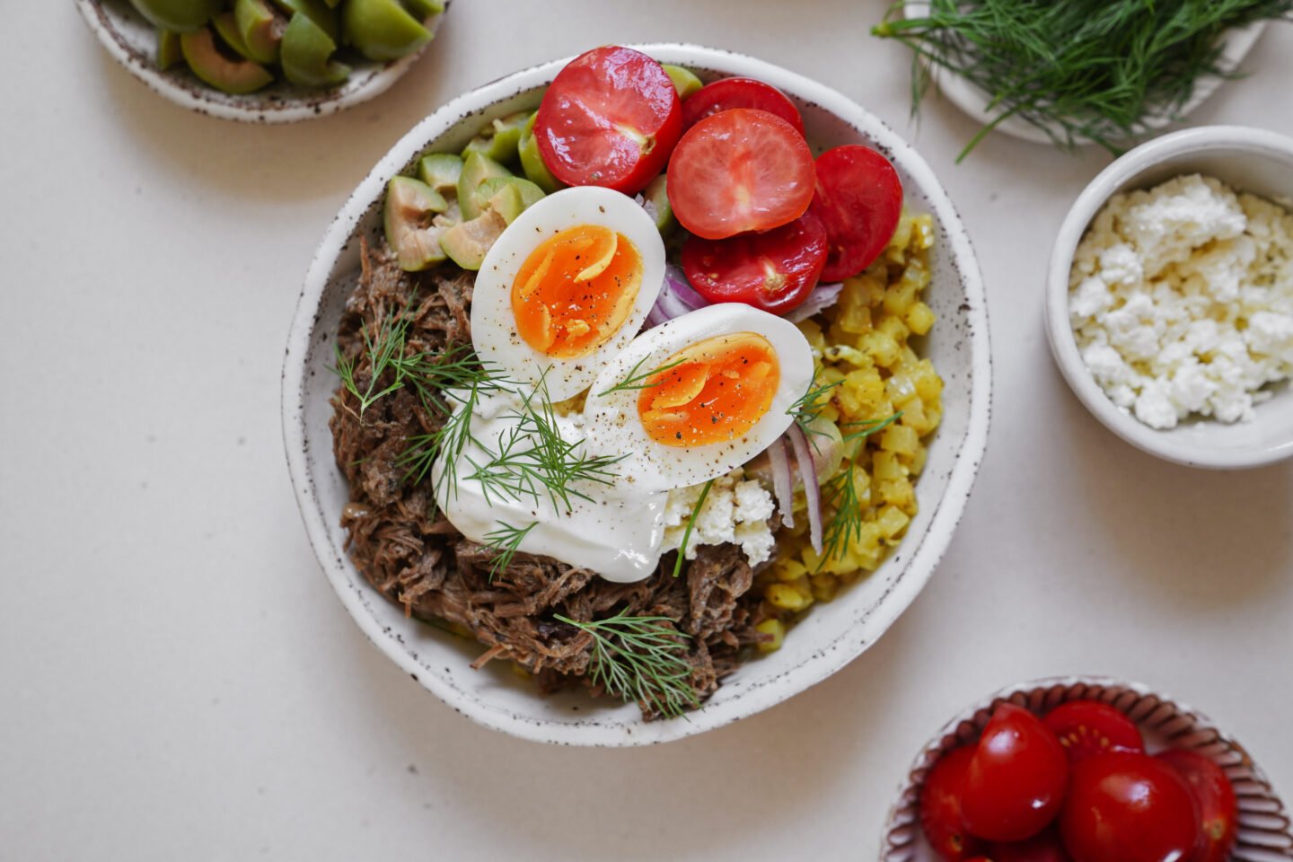 A bowl with shredded meat, sliced avocado, pickled onions, cherry tomatoes, yellow rice, dollops of white sauce, fresh dill, and two halved soft-boiled eggs. Sides of olives, ricotta, and cherry tomatoes surround it.