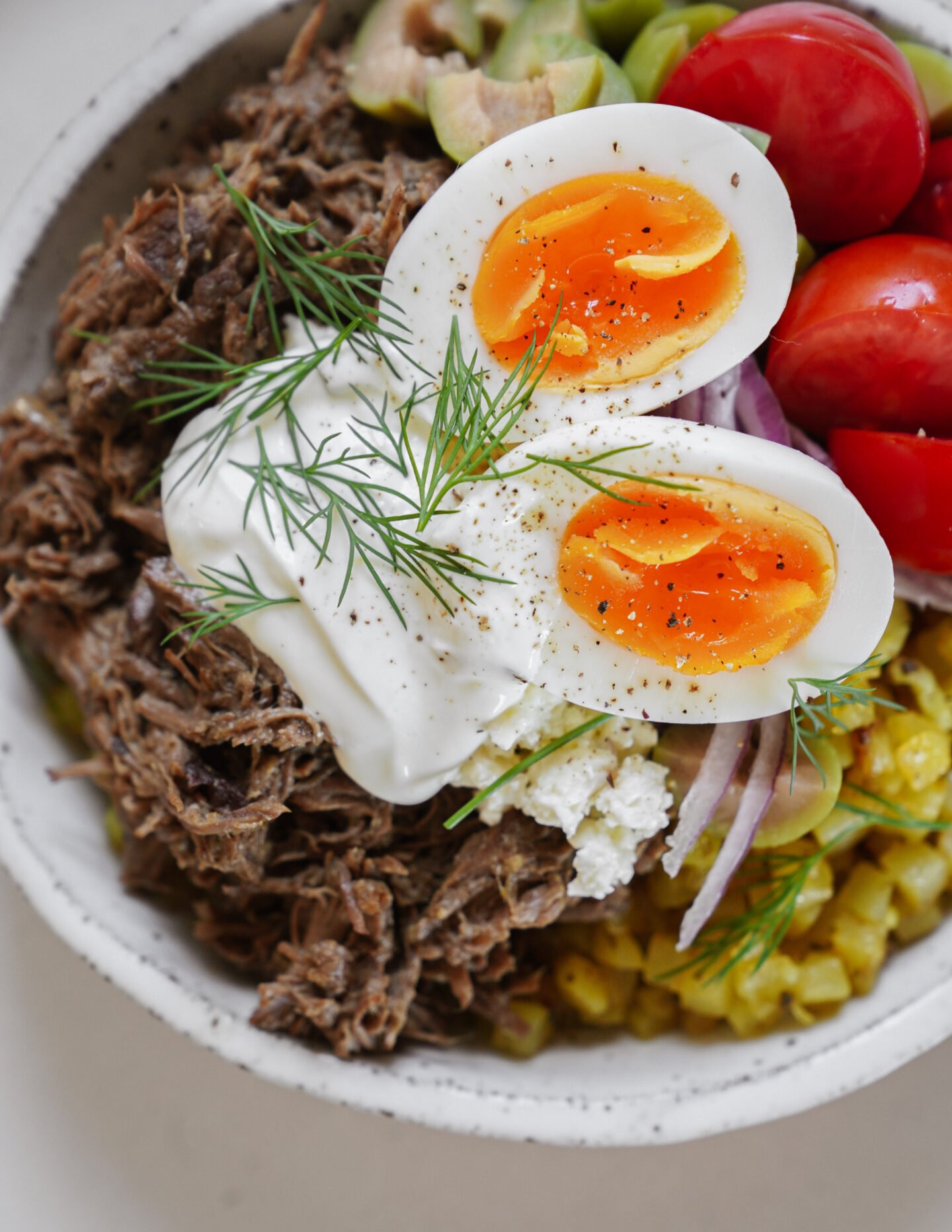 A bowl filled with shredded beef, halved soft-boiled eggs, cherry tomatoes, diced avocado, corn, red onions, sour cream, and fresh dill, all arranged in separate sections.