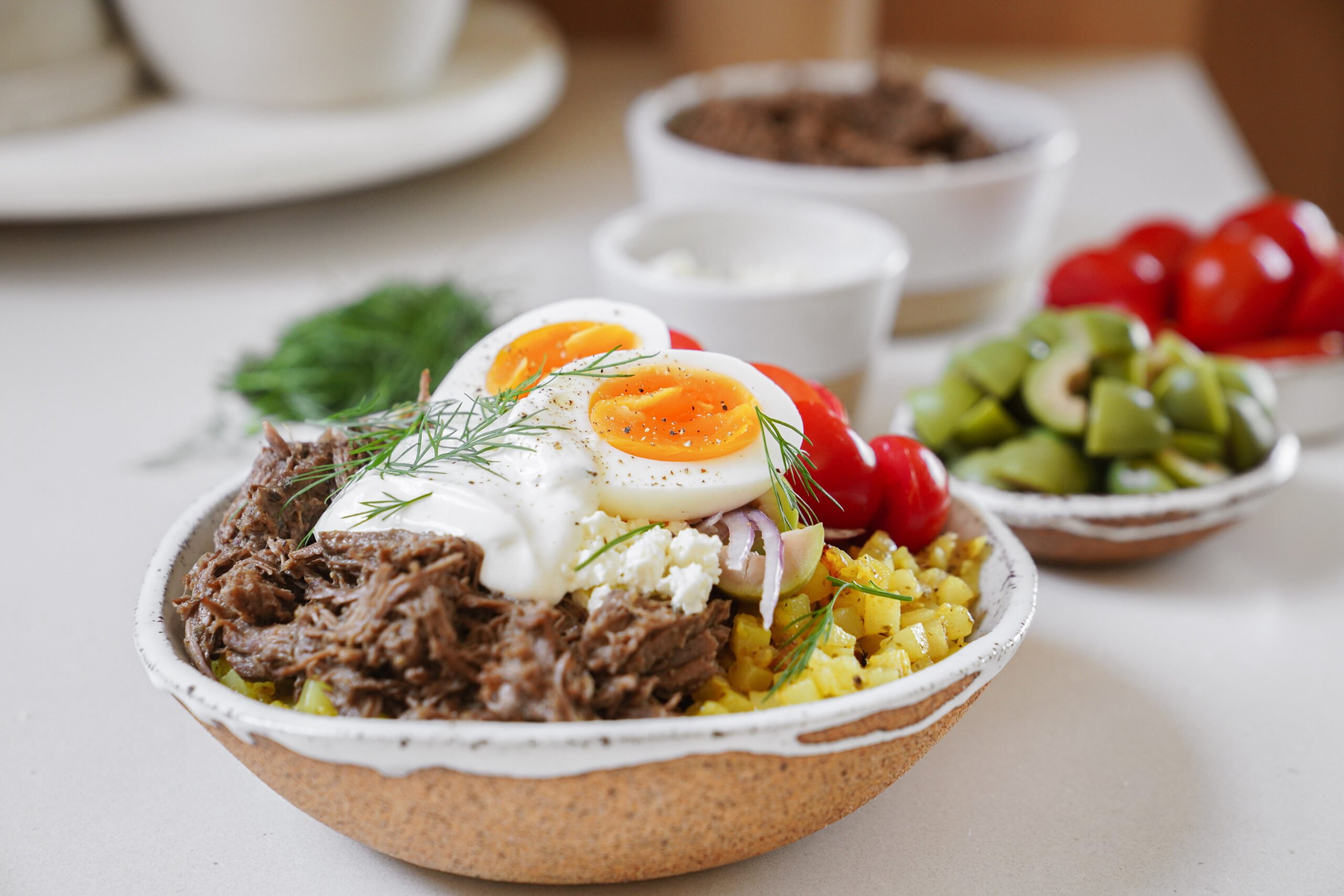 A bowl filled with shredded beef, sliced boiled egg, cherry tomatoes, olives, grains, and a dollop of yogurt, garnished with fresh dill, sits on a white surface with ingredients in bowls in the background.