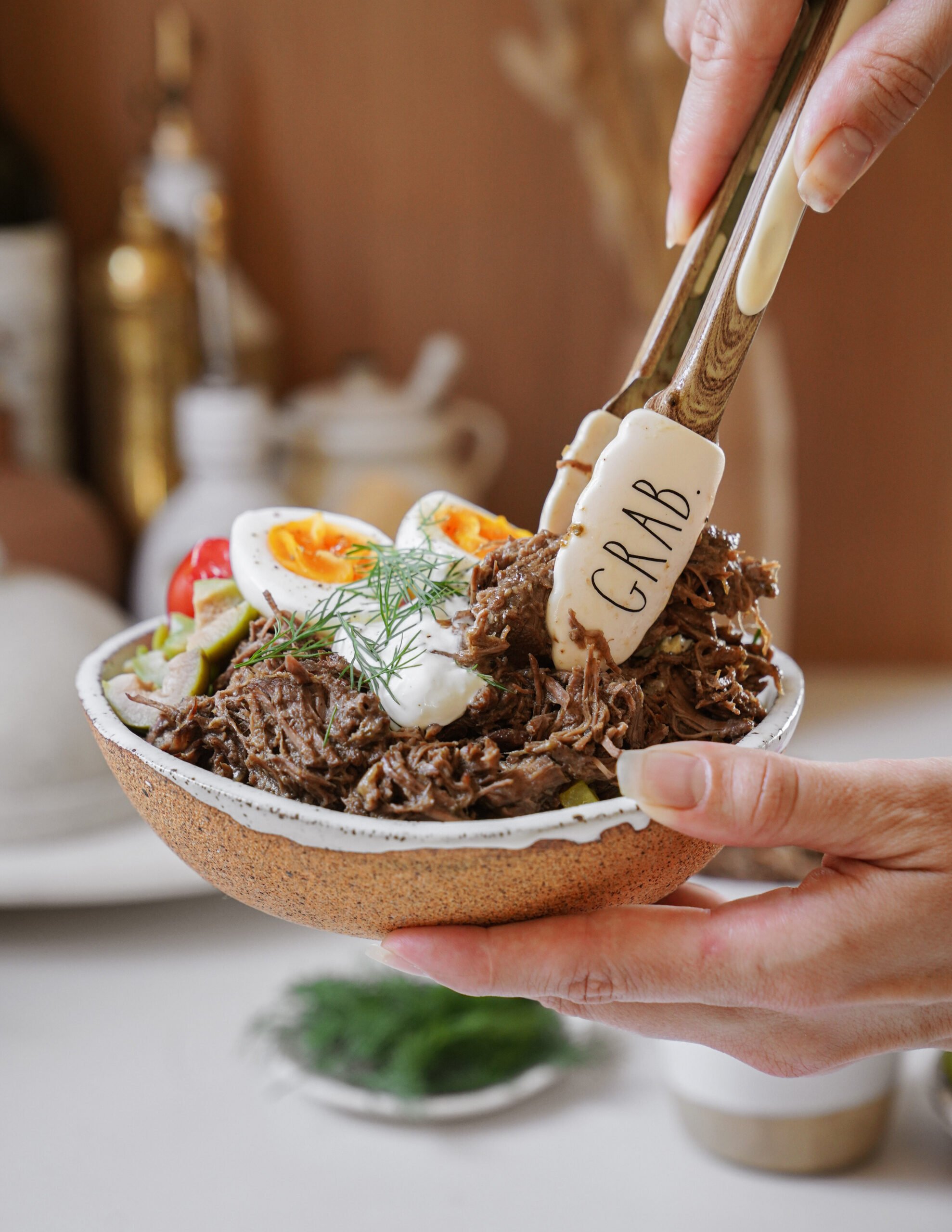 A hand holds a bowl filled with shredded beef, sliced boiled eggs, cherry tomatoes, dill, and greens, using tongs labeled GRAB to pick up the meat. The background is softly blurred.