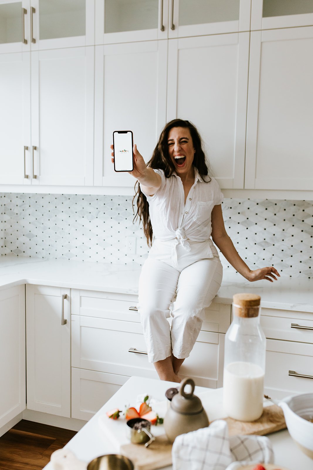 A woman in a white outfit sits on a kitchen counter, joyfully holding up her phone. The kitchen features white cabinets and a decorative backsplash. A table in the foreground has a carafe of milk and some kitchen items.