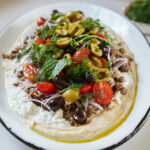 A platter of hummus topped with ground meat, cherry tomatoes, sliced olives, red onions, tzatziki, fresh dill, and mint leaves, drizzled with olive oil.