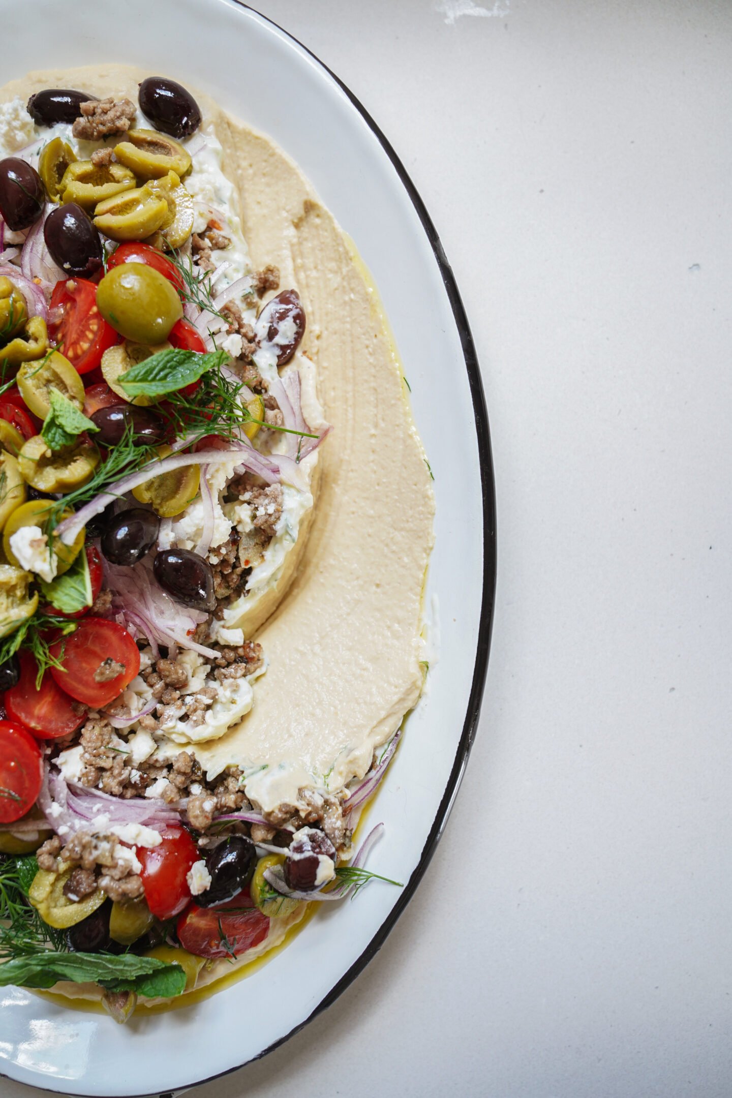 A white oval plate with creamy hummus topped with ground meat, sliced tomatoes, red onions, mixed olives, fresh herbs, and a drizzle of olive oil, arranged artfully on a light surface.