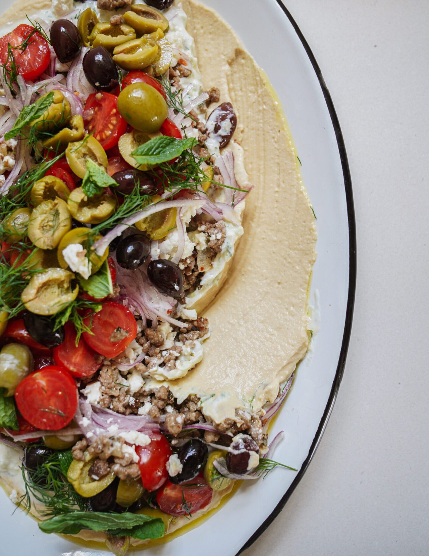 A platter topped with creamy hummus, sliced tomatoes, olives, red onions, ground meat, crumbled cheese, and fresh herbs, arranged artfully on a white plate with a black rim.