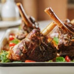 Close-up of roasted lamb shanks garnished with fresh herbs and cherry tomatoes, served in a white rectangular dish. The meat appears tender and well-seasoned.