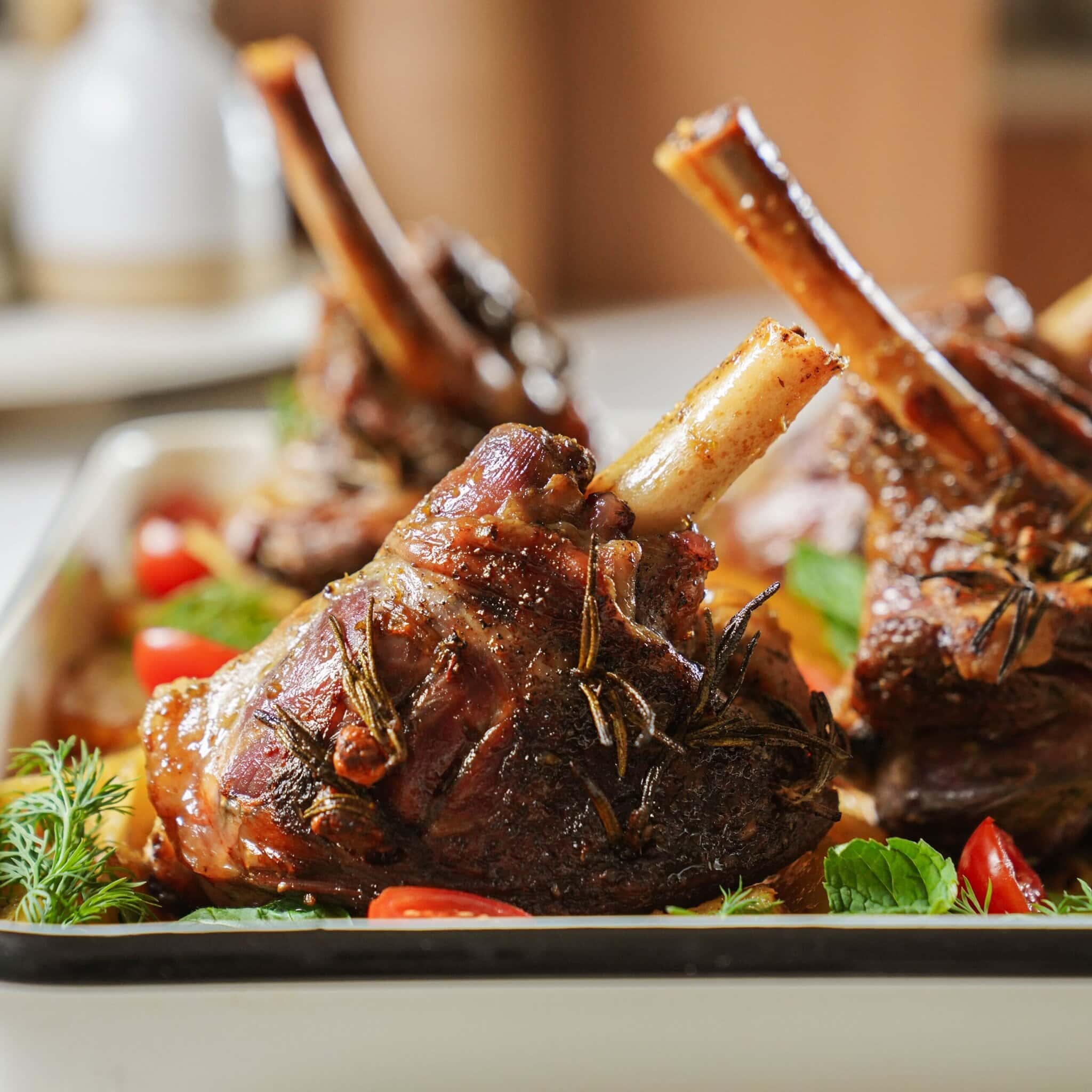 Close-up of roasted lamb shanks garnished with fresh herbs and cherry tomatoes, served in a white rectangular dish. The meat appears tender and well-seasoned.
