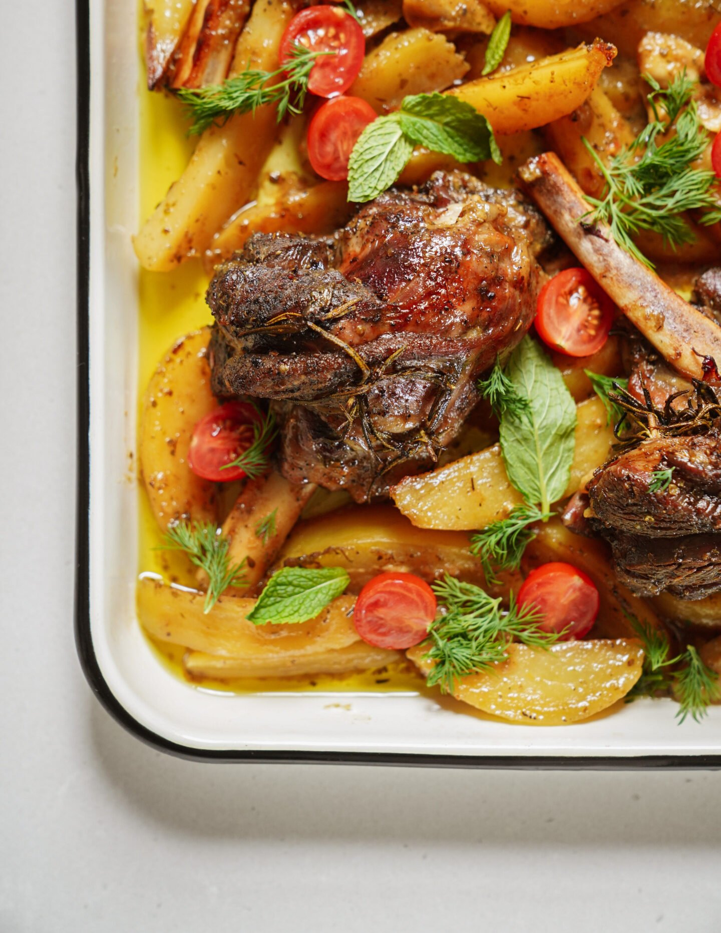 A roasted lamb shank with golden potato wedges, cherry tomato halves, and fresh herbs in a white baking dish.