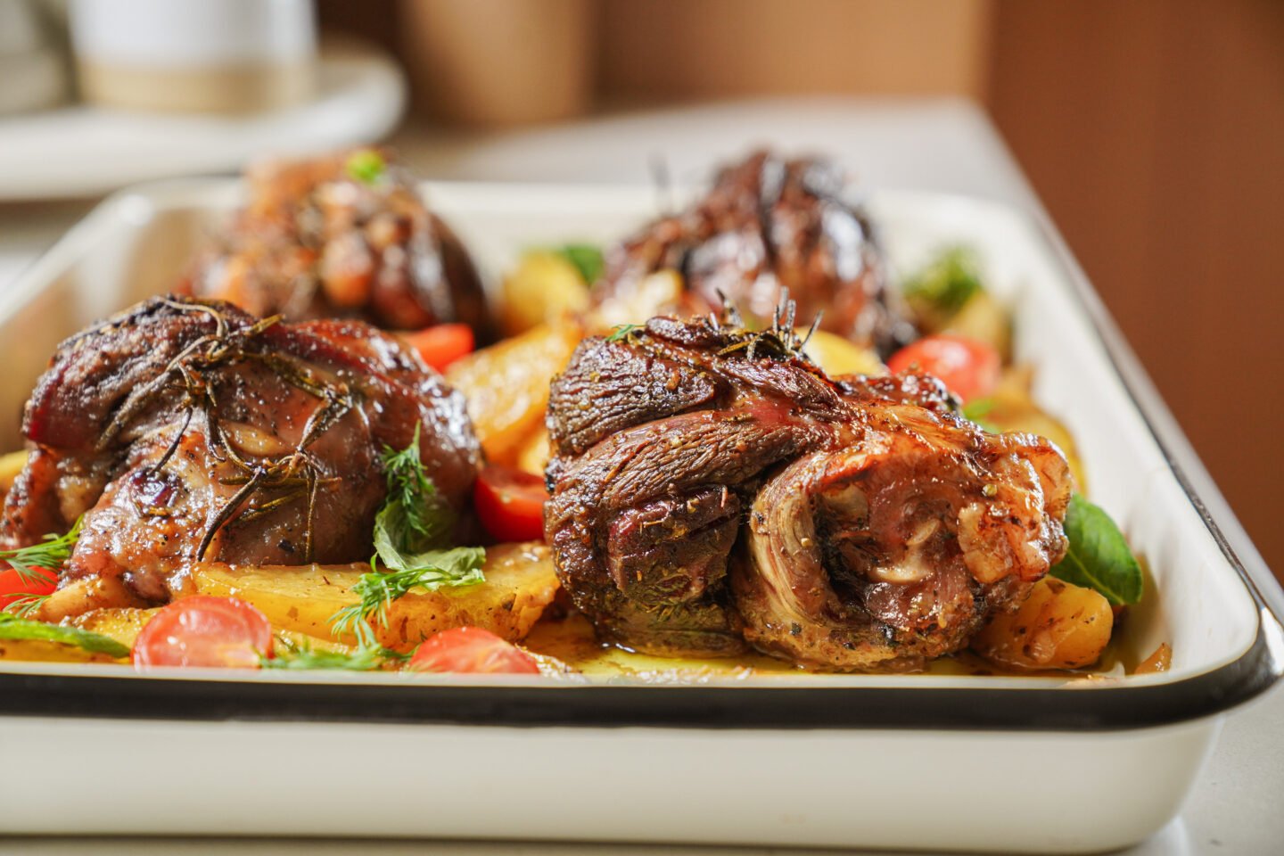 A close-up of roasted lamb shanks garnished with herbs, served on a tray with golden potatoes and cherry tomatoes. The dish looks juicy and flavorful, with a crispy, caramelized exterior.