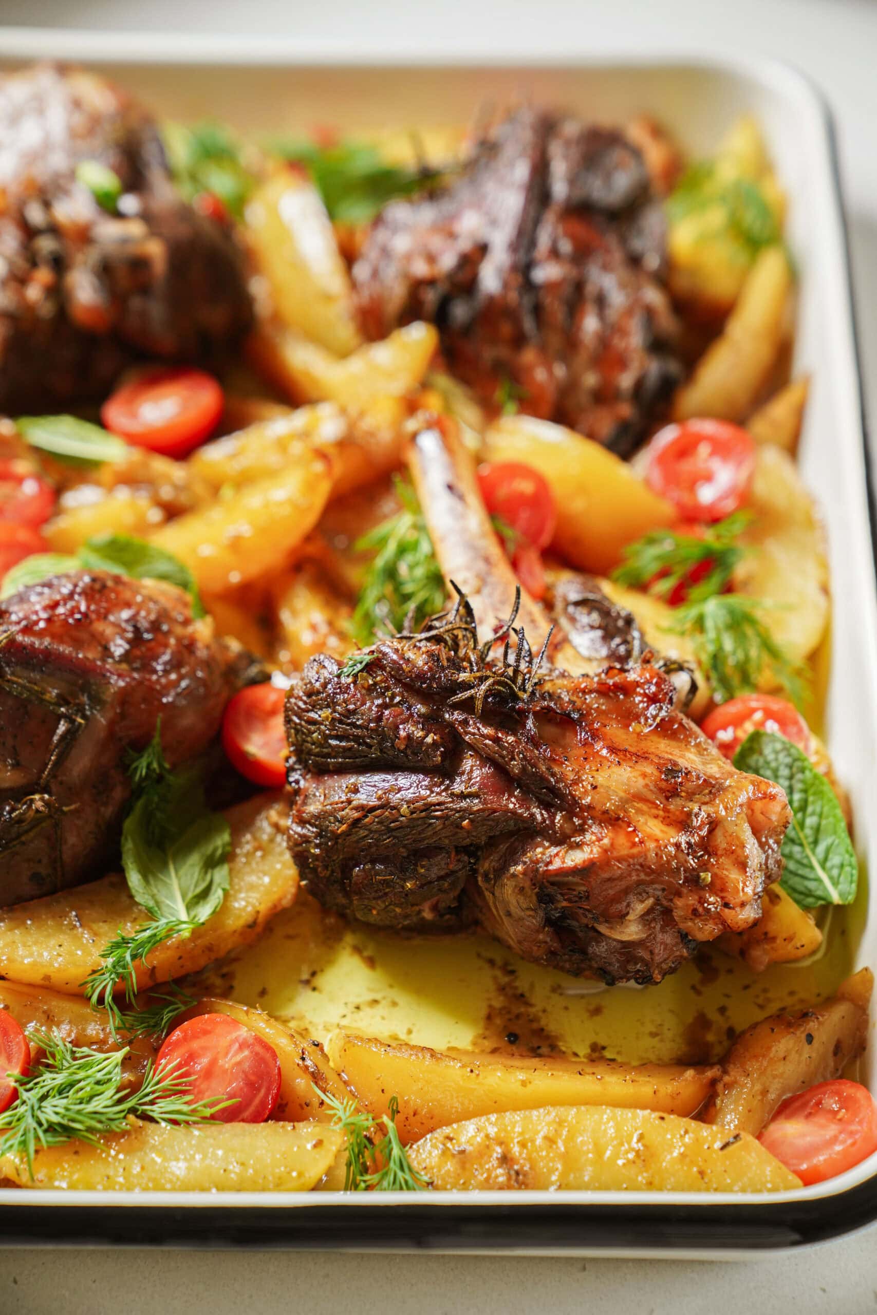Roasted lamb shanks with golden potato wedges, cherry tomatoes, and fresh herbs on a baking tray. The dish is colorful, well-seasoned, and has a rustic, homemade appearance.