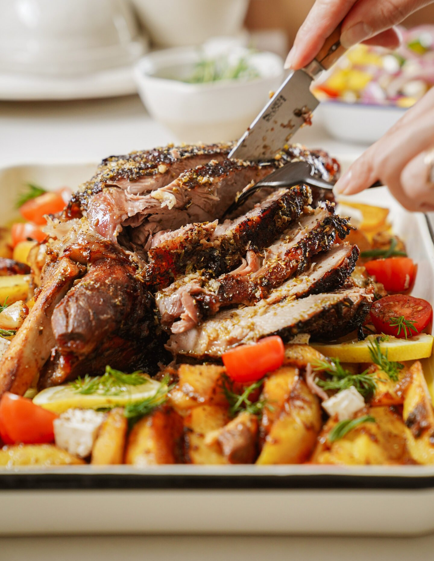 A well-seasoned roast, being sliced with a knife. The roast rests on a bed of roasted potatoes and garnished with cherry tomatoes and herbs. A bowl of salad and some dinnerware are visible in the blurred background.