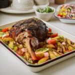 Roasted lamb shank on a bed of sliced potatoes in a rectangular dish, garnished with cherry tomatoes, herbs, and cheese. A salad with peppers and onions is visible in the background.