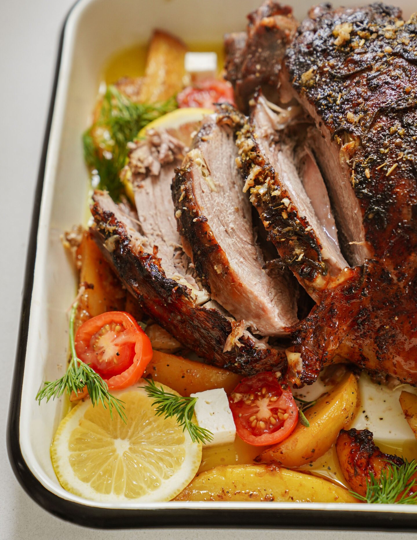 A roasted, seasoned meat dish with slices on display. It rests on a bed of potato wedges, cherry tomatoes, lemon slices, and garnished with fresh dill in a white baking dish.