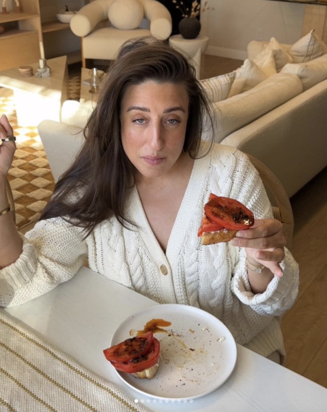 A person with long dark hair is sitting at a table, holding a piece of toast topped with sliced tomatoes. They are wearing a knitted cream sweater. The table has a white plate with toast and a striped tablecloth. The background shows a cozy living room.