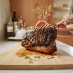 A person holds a cooked, herb-crusted rack of ribs on a wooden cutting board in a kitchen. The ribs are garnished with fresh herbs. Bottles and kitchenware are visible in the blurred background.