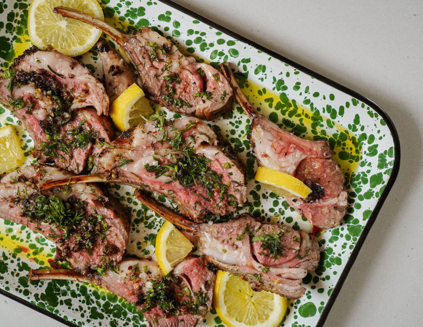 Grilled lamb chops garnished with herbs are placed on a speckled green and white platter. Lemon wedges are scattered around for garnish.
