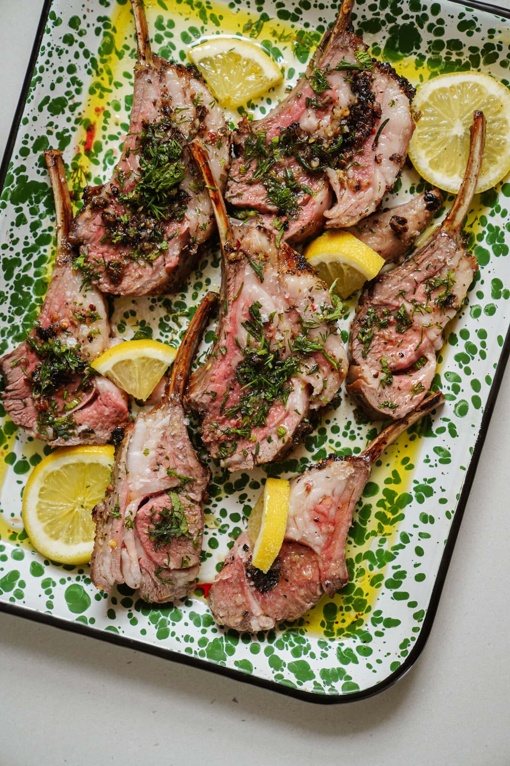 Grilled lamb chops garnished with herbs are arranged on a speckled green and white platter. Slices of lemon are scattered among the chops, adding a touch of citrus to the presentation.