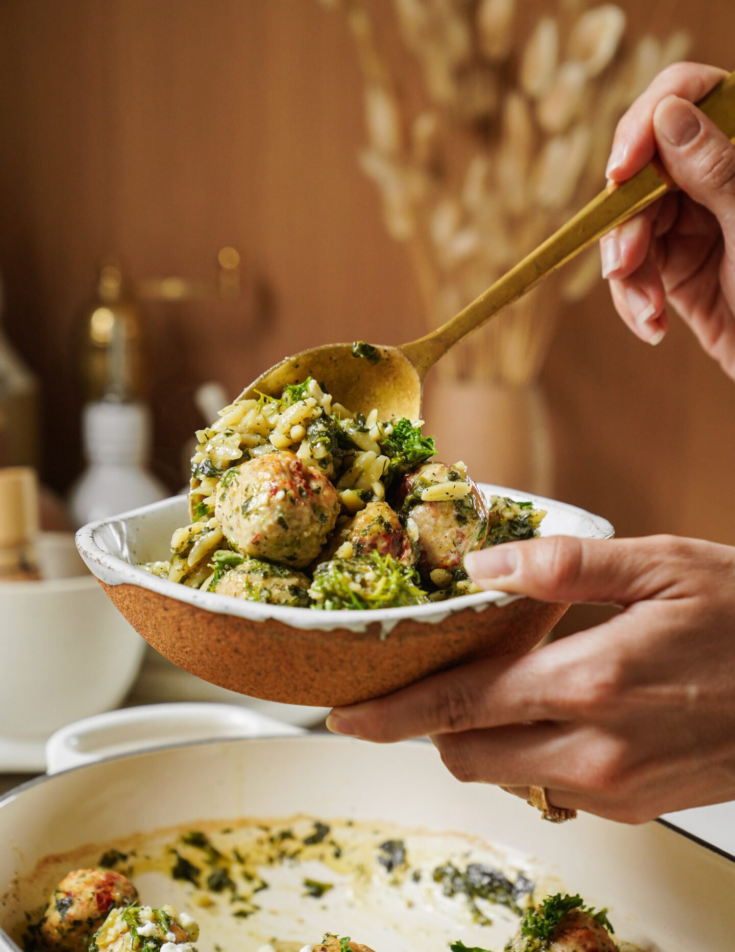 A person serves a bowl of pasta with meatballs, broccoli, and herbs from a white casserole dish using a golden spoon, with a blurred kitchen background.